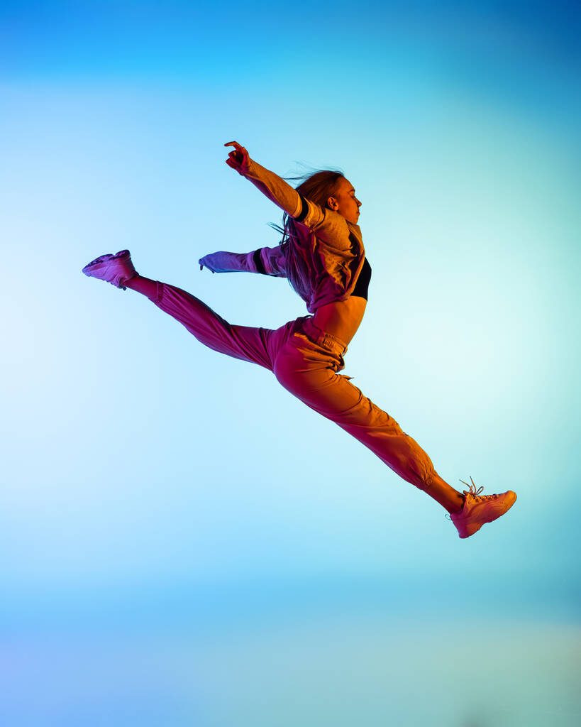 Jedna młoda sportowa dziewczyna tańcząca hip-hop w stylowych ciuchach na kolorowym tle w sali tanecznej w neonowym świetle. Kultura młodzieżowa, ruch, styl i moda, akcja. - Zdjęcie, obraz