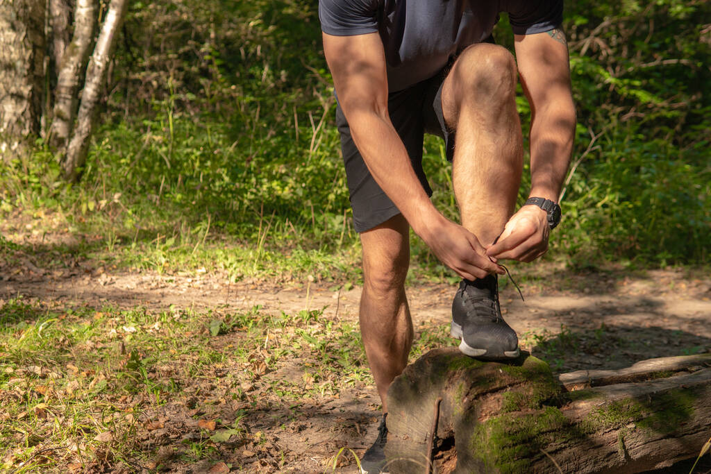 De sporter knoopt de veters van zijn benen in sneakers van dichtbij de atleet loopt in het park buiten, rond het bos, eiken bomen groen gras jonge duurzame atleet lopen atleet fitness - Foto, afbeelding