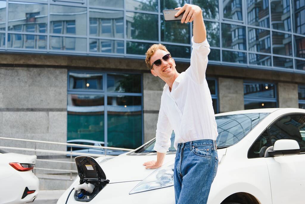 Portrait de jeune homme européen beau, faisant selfie photo sur son smartphone tout en s'appuyant sur sa voiture électrique, rechargeant la batterie à la centrale électrique de la ville après avoir acheté des produits frsh dans le centre commercial - Photo, image