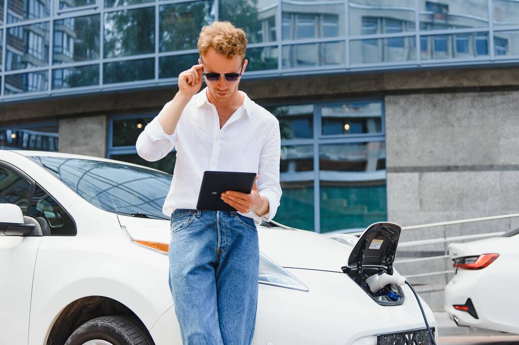 Obchodní muž stojí v blízkosti nabíjení elektrického auta a pomocí tabletu na ulici. - Fotografie, Obrázek