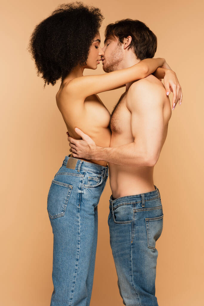 vista lateral de casal interracial apaixonado em jeans apenas abraçando e beijando isolado no bege - Foto, Imagem