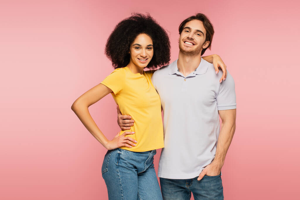 junges und fröhliches interrassisches Paar blickt in die Kamera, während es sich isoliert auf rosa umarmt - Foto, Bild