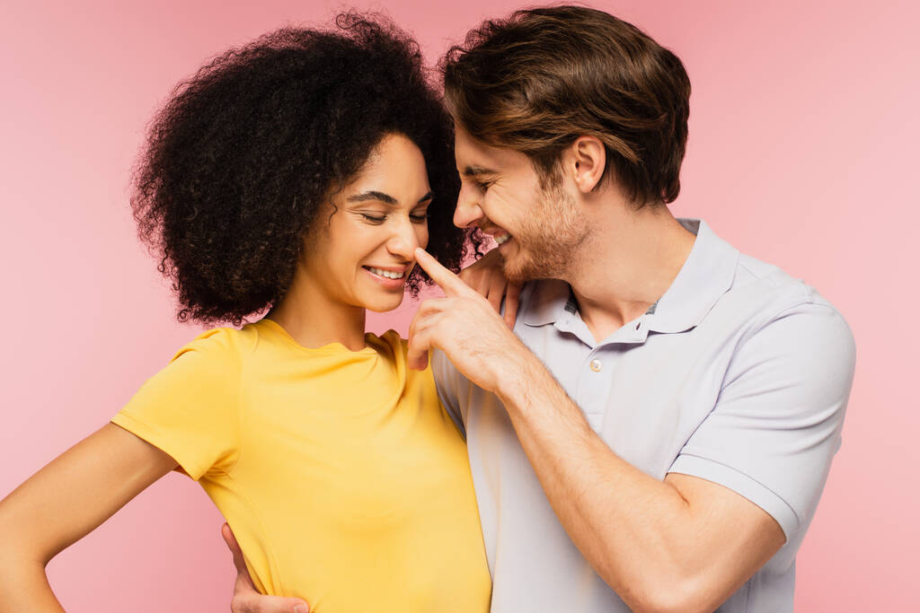 homme joyeux étreignant heureuse petite amie hispanique tout en touchant son nez isolé sur rose - Photo, image
