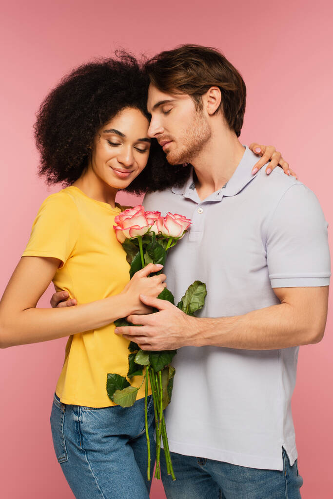 щаслива міжрасова пара з букетом з троянд, що обіймаються закритими очима ізольовані на рожевому
 - Фото, зображення