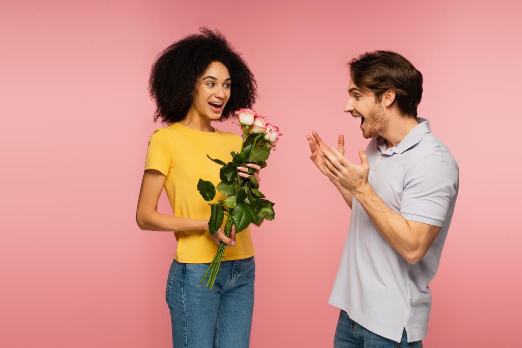 šťastný latinsky žena drží kytice růží v blízkosti ohromený muž ukazuje wow gesto izolované na růžové - Fotografie, Obrázek