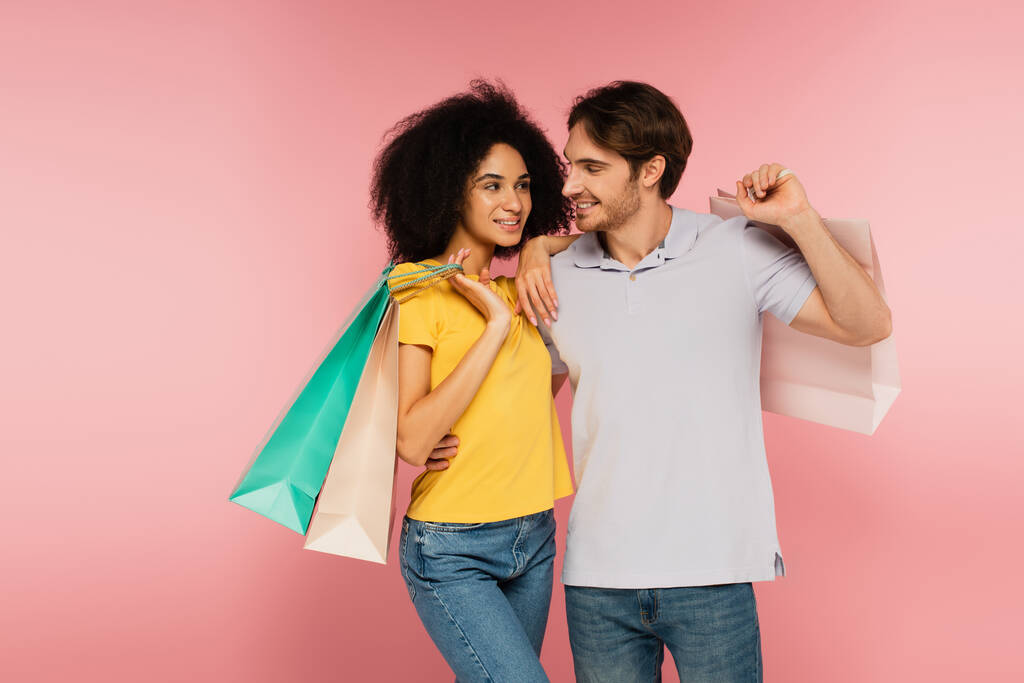 ευτυχισμένο διαφυλετικό ζευγάρι με τσάντες ψώνια κοιτάζοντας ο ένας τον άλλο απομονωμένο σε ροζ - Φωτογραφία, εικόνα