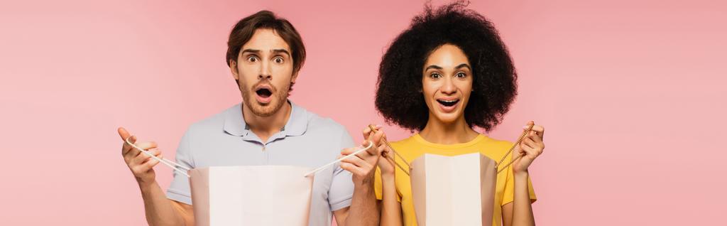 thrilled interracial couple with shopping bags looking at camera isolated on pink, banner - Photo, Image