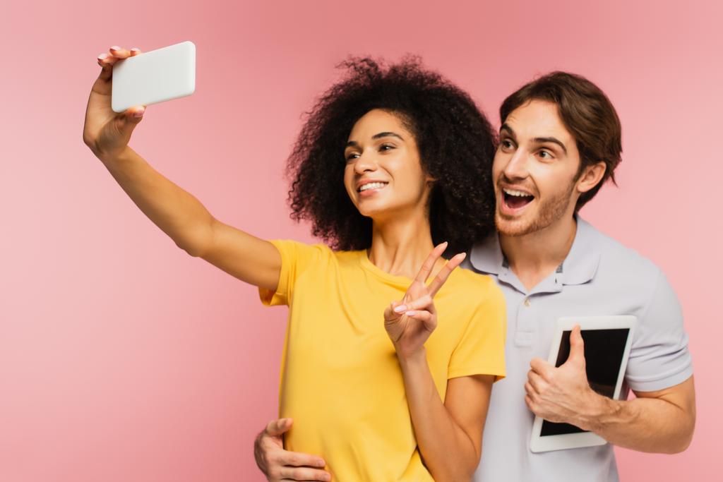 amazed man with digital tablet hugging hispanic girlfriend showing victory sign while taking selfie isolated on pink - Photo, Image
