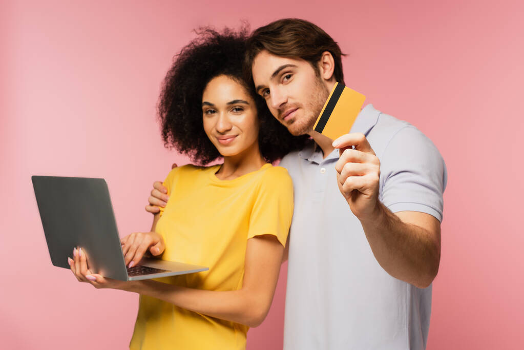 homme montrant carte de crédit et embrassant petite amie hispanique en utilisant ordinateur portable isolé sur rose - Photo, image