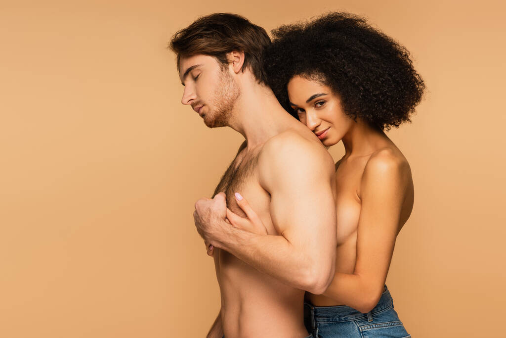 sensuale e topless donna ispanica abbracciando uomo senza maglietta in piedi con gli occhi chiusi isolati su beige - Foto, immagini