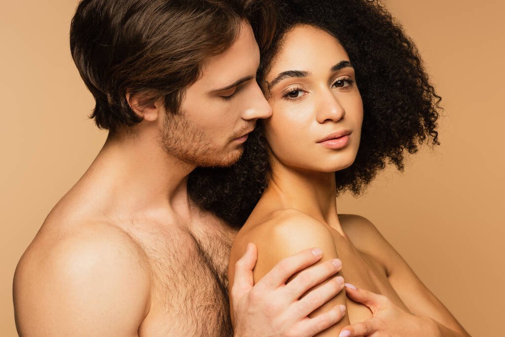hombre sin camisa con los ojos cerrados abrazando mujer hispana sexy mirando la cámara aislada en beige - Foto, imagen