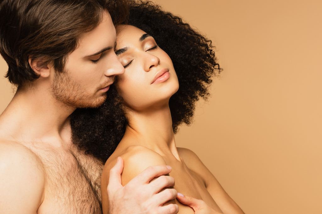 shirtloze man knuffelen sensuele latino vrouw staan met gesloten ogen geïsoleerd op beige - Foto, afbeelding