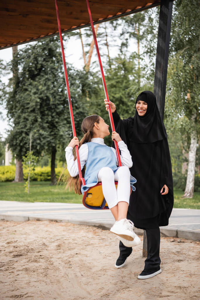 Salıncakta sallanan kızın yanında duran gülümseyen Müslüman anne.  - Fotoğraf, Görsel