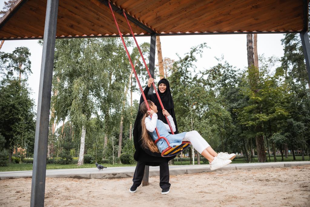 Vrolijke moslim moeder in hijab staan in de buurt dochter op swing in park  - Foto, afbeelding
