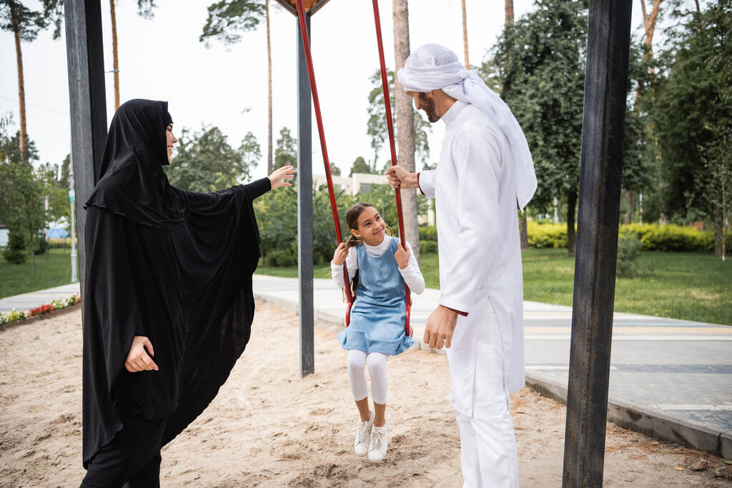 Улыбающийся ребенок смотрит на арабских родителей, размахивая в парке  - Фото, изображение