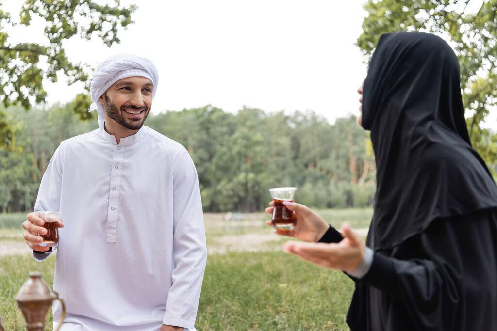 uśmiechnięty arabski mężczyzna trzymając szklankę herbaty w pobliżu zamazanej żony w parku  - Zdjęcie, obraz
