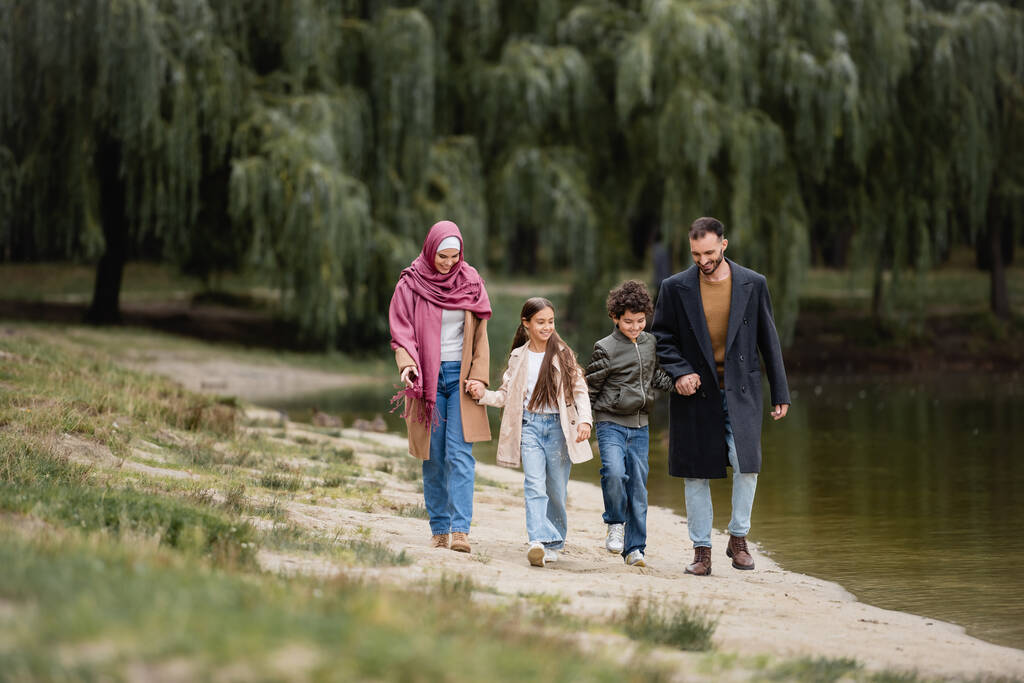 Hymyilevät muslimilapset kävelemässä lähellä vanhempia ja järveä puistossa  - Valokuva, kuva