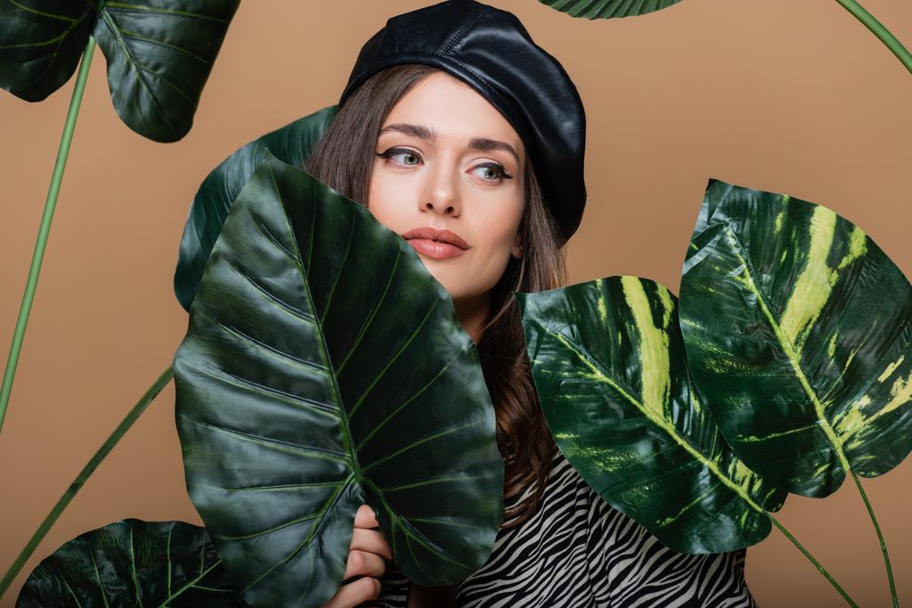 mladá žena v koženém baretu v blízkosti tropických listí izolovaných na béžové  - Fotografie, Obrázek