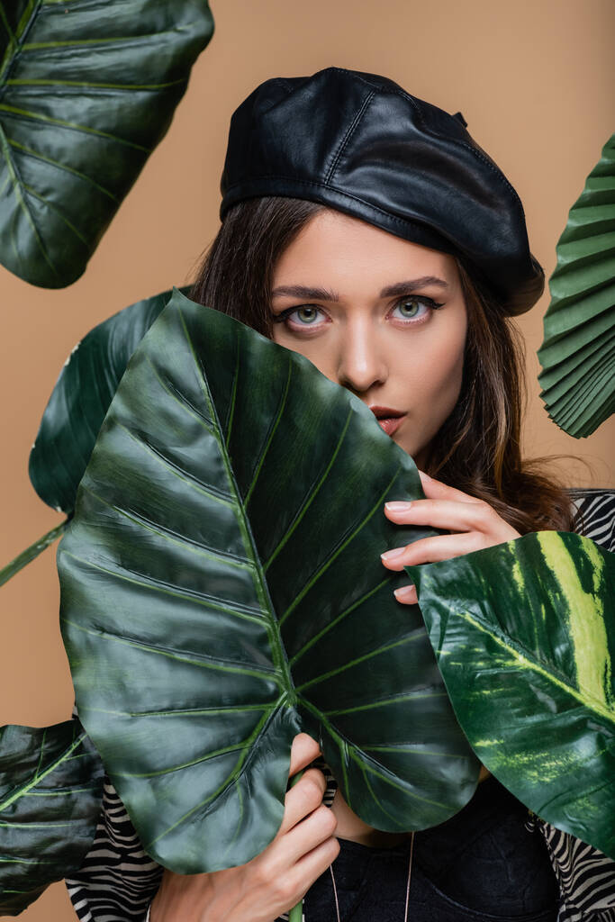 giovane donna in berretto nero guardando la macchina fotografica attraverso foglie tropicali verdi isolate sul beige  - Foto, immagini