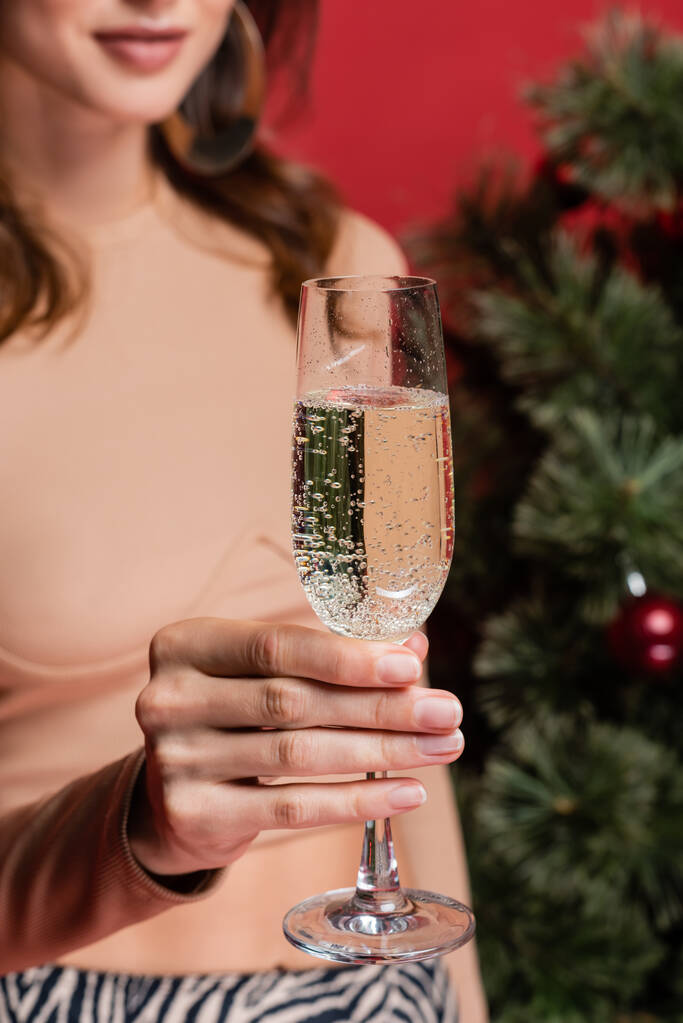 赤で隔絶されたクリスマスツリーの近くにシャンパンのグラスを持っているぼやけた女性のクロップドビュー  - 写真・画像