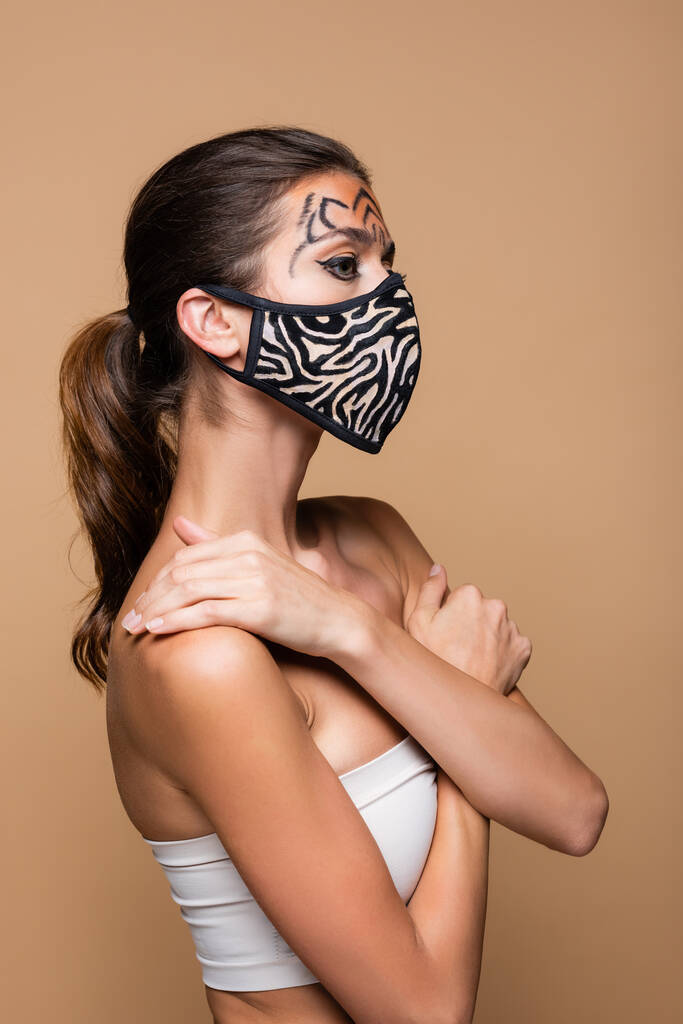 mujer joven con maquillaje de tigre y máscara protectora de impresión animal posando aislado en beige - Foto, Imagen