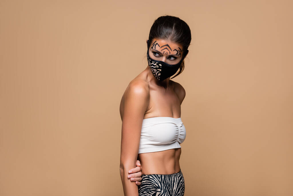 γυναίκα με τίγρη μακιγιάζ και ζώων εκτύπωσης προστατευτική μάσκα κοιτάζοντας μακριά απομονώνονται σε μπεζ - Φωτογραφία, εικόνα