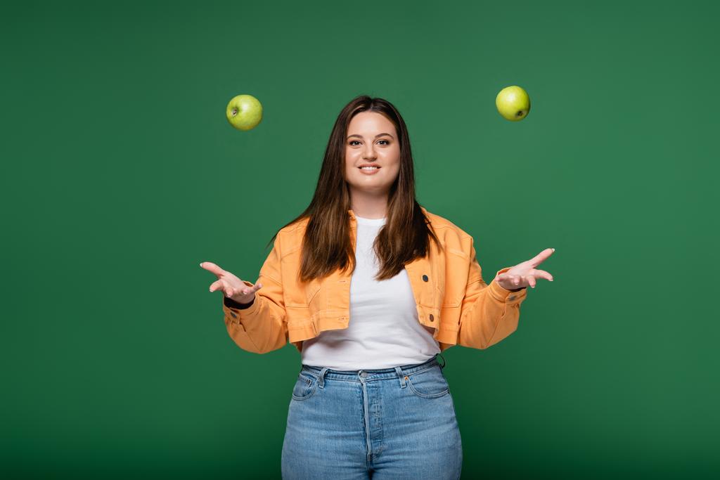 Glimlachende vrouw met overgewicht gooien appels geïsoleerd op groen  - Foto, afbeelding