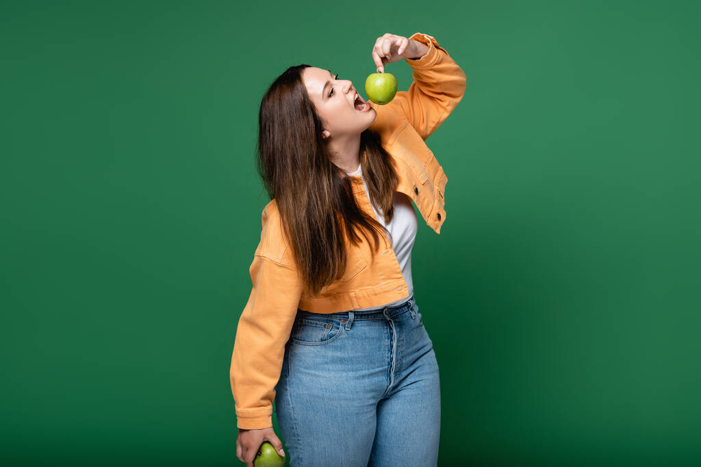 Joven más tamaño mujer sosteniendo manzana cerca de boca abierta aislado en verde  - Foto, imagen