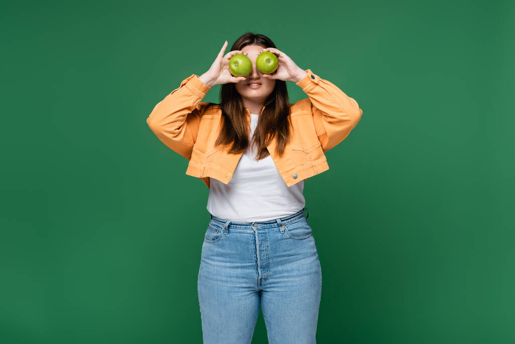 Uśmiechnięta kobieta z nadwagą trzymająca zielone jabłka w pobliżu oczu odizolowanych na zielono  - Zdjęcie, obraz