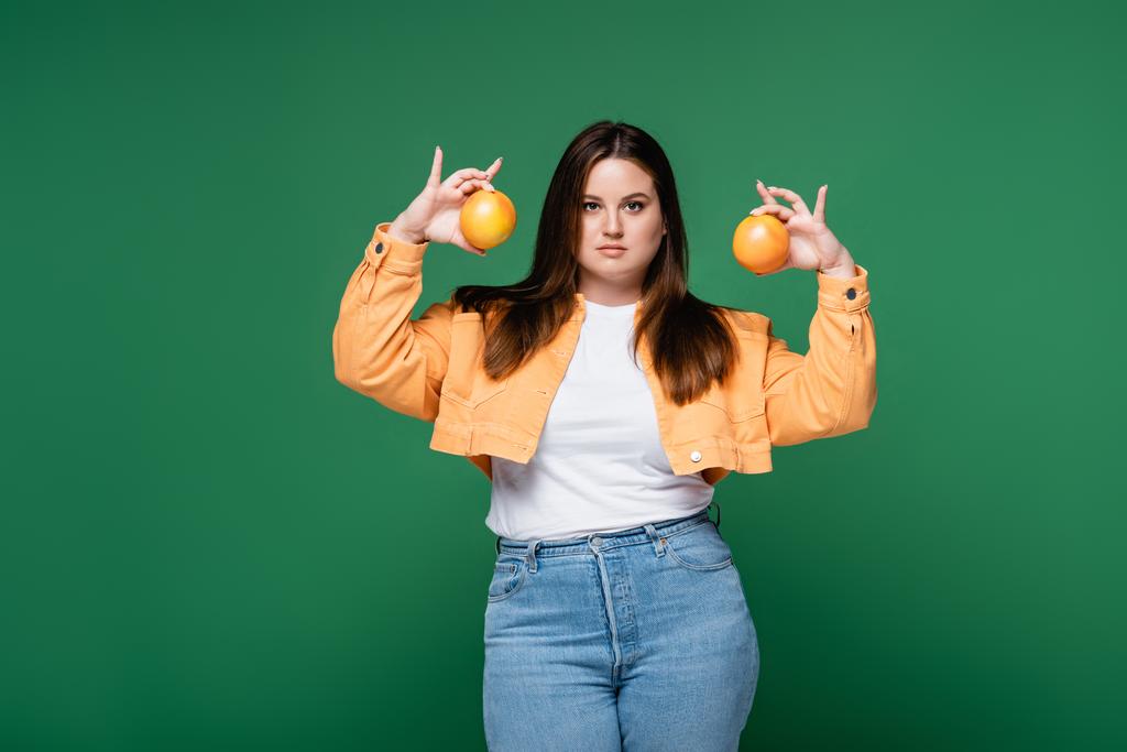 Νεαρή γυναίκα με υπέρβαρα πορτοκάλια που διατηρούνται απομονωμένα στο πράσινο  - Φωτογραφία, εικόνα