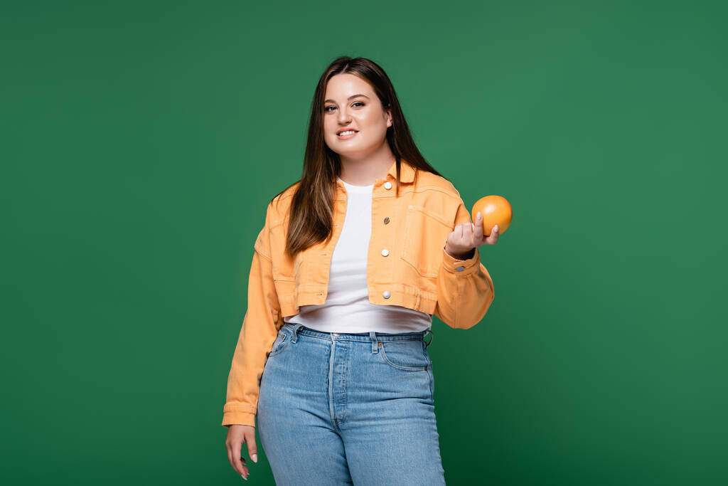 Positive woman with overweight holding orange and smiling at camera isolated on green  - Photo, Image