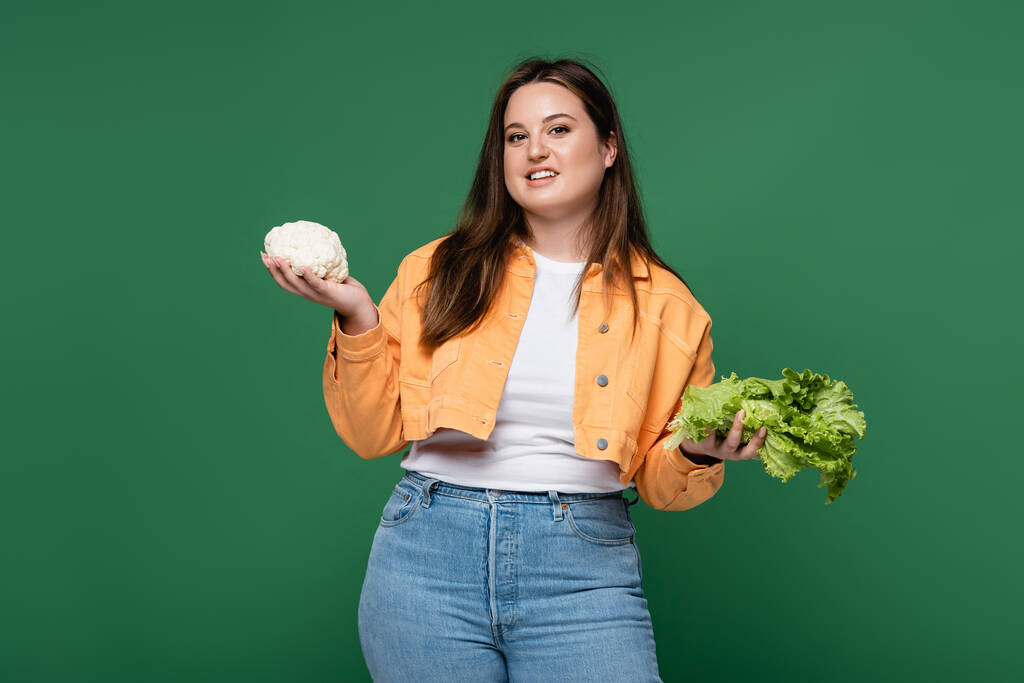 Улыбающееся тело позитивная женщина держит салат и цветную капусту изолированы на зеленом  - Фото, изображение