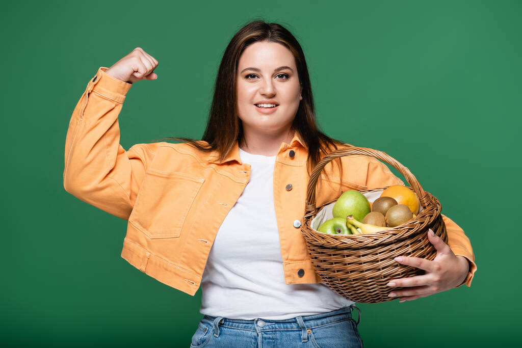 Mosolygó nő túlsúlyos bemutató izmok és gazdaság kosár gyümölcsök elszigetelt zöld  - Fotó, kép