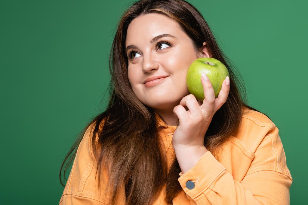 Cheerful brunette woman with overweight holding fresh apple isolated on green  - Photo, Image