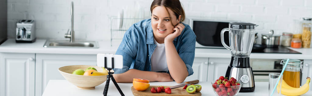 Положительная женщина с избыточным весом глядя на смартфон рядом фрукты на кухонном столе, баннер  - Фото, изображение
