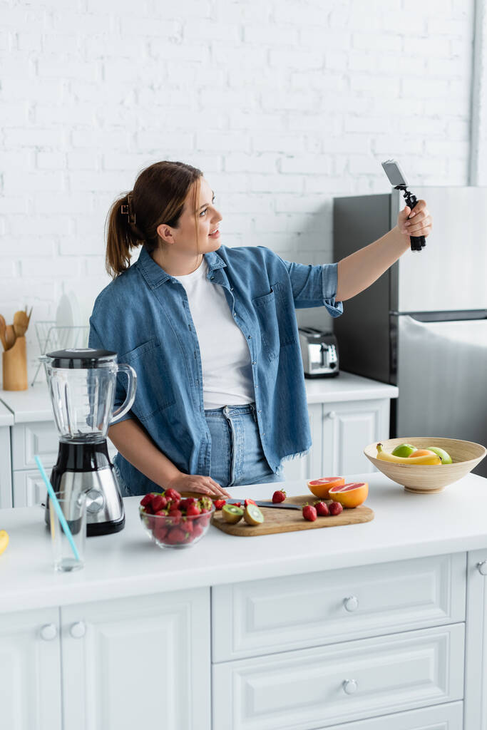 Lächelnder Blogger mit Übergewicht nutzt Smartphone in der Nähe von Mixer und Obst in der Küche  - Foto, Bild