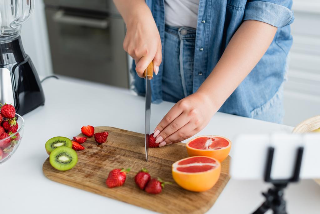 Vista recortada de la mujer joven cortando frutas frescas cerca de licuadora y teléfono inteligente borroso en la cocina  - Foto, Imagen
