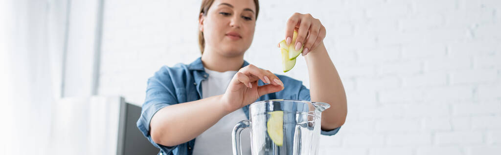 Aşırı kilolu bulanık bir kadın mutfaktaki blender 'a elma dilimleri koyuyor.  - Fotoğraf, Görsel