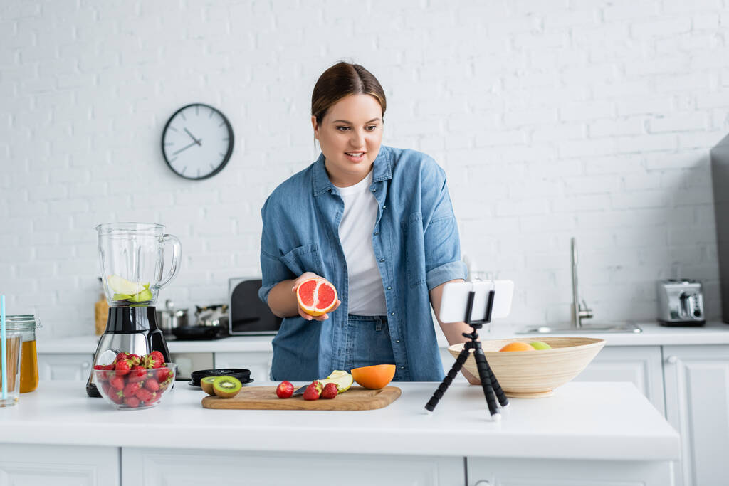 Souriant femme plus taille tenant pamplemousse près de fruits et smartphone dans la cuisine  - Photo, image
