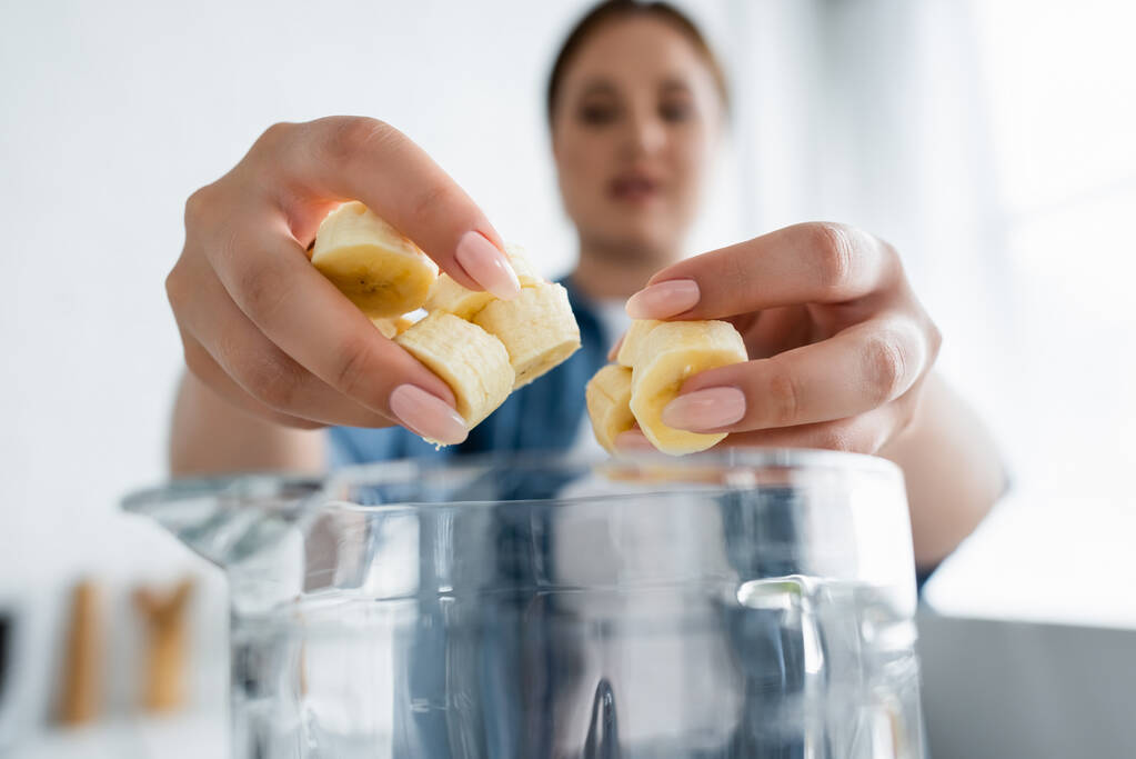 Donna offuscata che tiene la banana affettata vicino al frullatore in cucina  - Foto, immagini