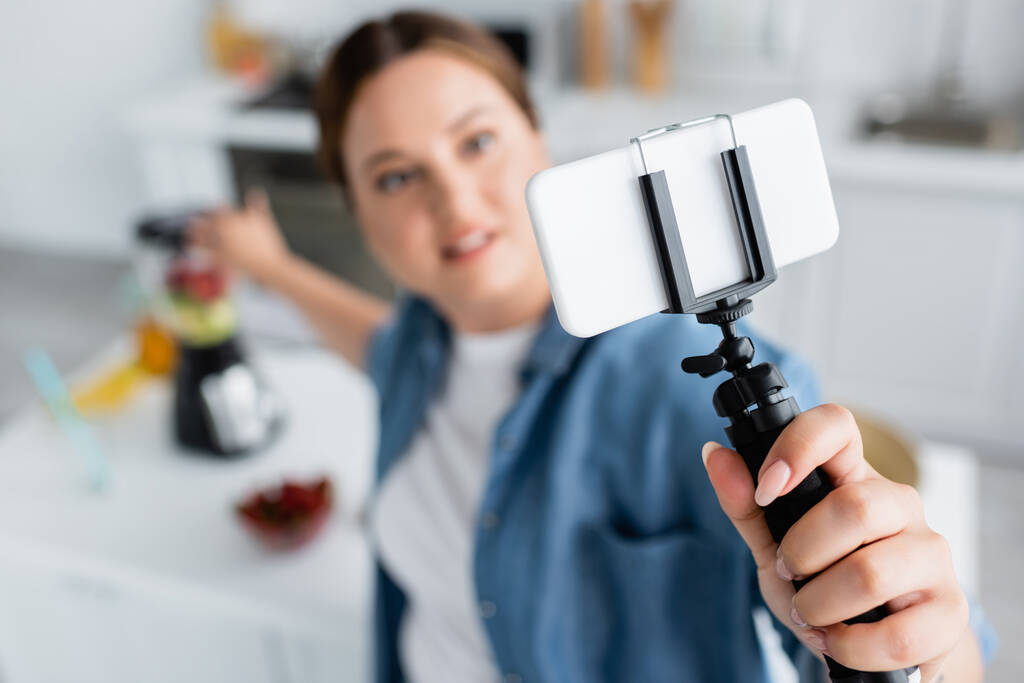 Vista de ángulo alto del teléfono inteligente en el trípode en la mano de la mujer borrosa más tamaño en la cocina  - Foto, Imagen