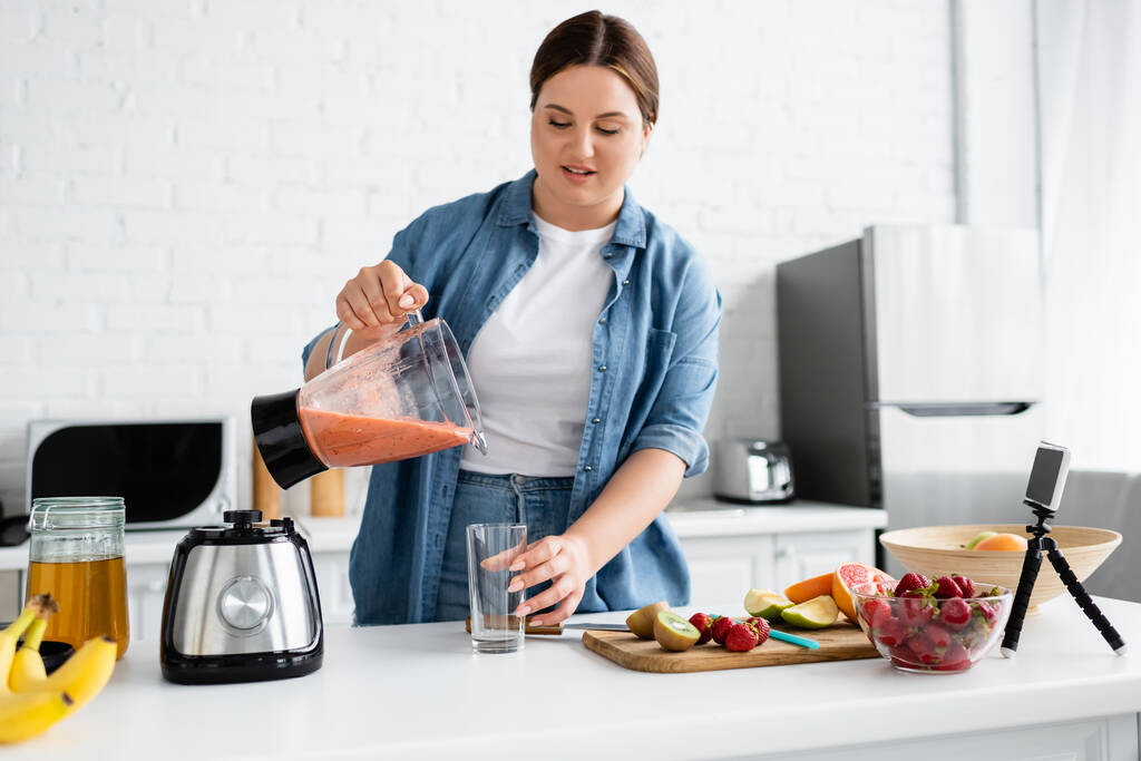 Young woman with overweight pouring fruit smoothie near smartphone in kitchen  - Photo, Image