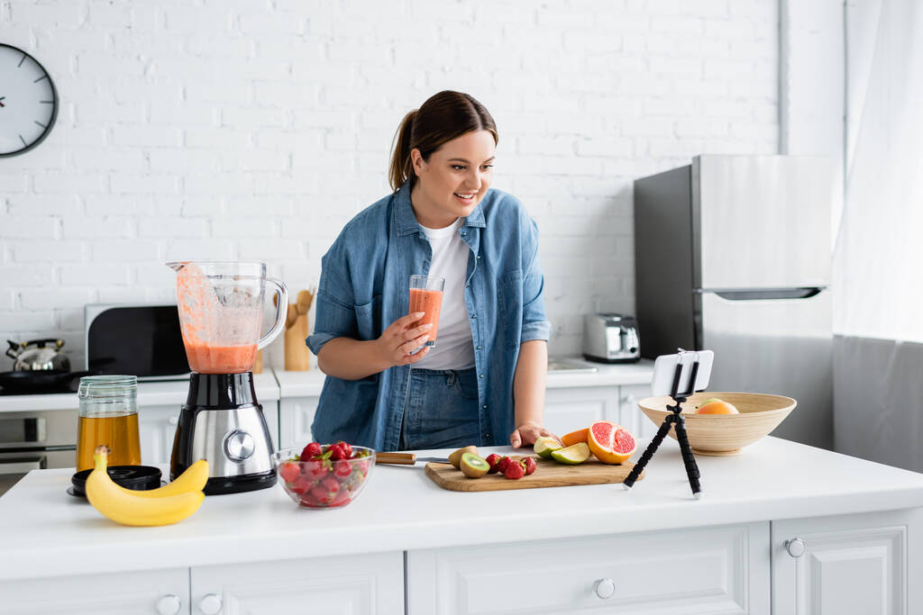 Alegre mujer de tamaño grande sosteniendo un vaso de batido cerca de frutas y teléfonos inteligentes en la cocina  - Foto, imagen
