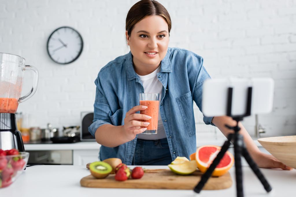 Plus-Size-Frau hält Glas Smoothie neben verschwommenem Handy auf Stativ in Küche  - Foto, Bild