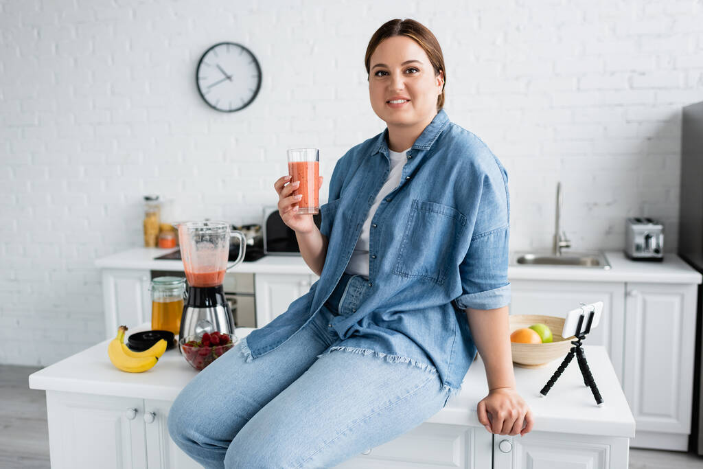 Usmívání plus velikost žena drží sklenici smoothie v blízkosti ovoce a smartphone na kuchyňském stole  - Fotografie, Obrázek