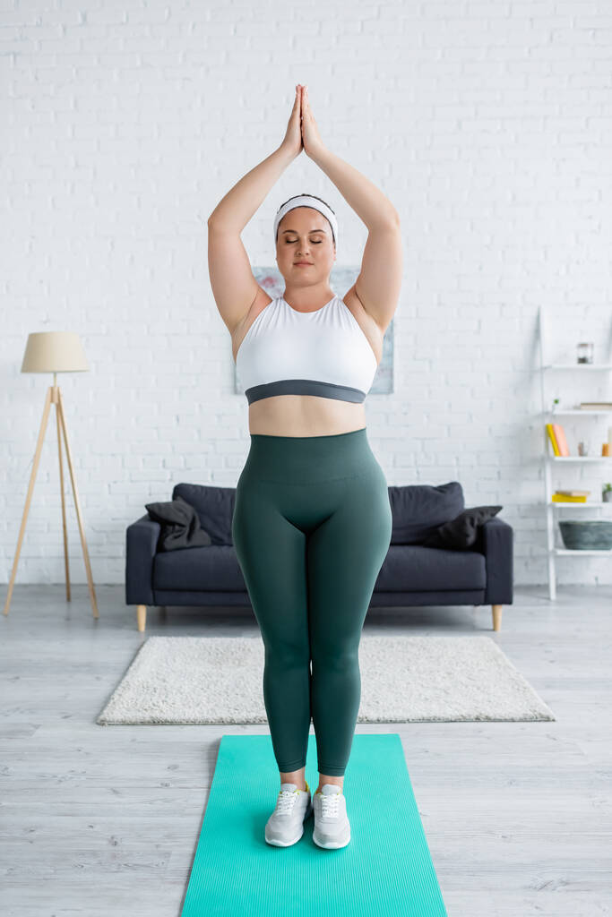 Plus size vrouw in sportkleding staande met biddende handen op fitness mat  - Foto, afbeelding