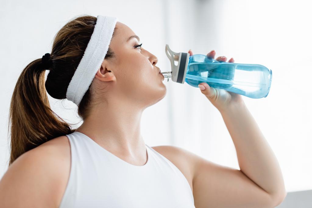 Seitenansicht einer Plus-Size-Frau in Sportbekleidung, die zu Hause Wasser trinkt  - Foto, Bild
