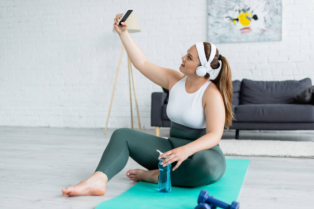 Вид сбоку улыбающейся plus size женщины в наушниках, делающей селфи на смартфоне рядом со спортивной бутылкой на фитнес-коврике  - Фото, изображение