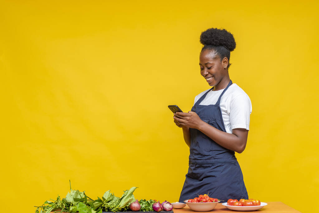 Una chef africana revisando su teléfono mientras prepara comida frente a una pared amarilla - Foto, Imagen