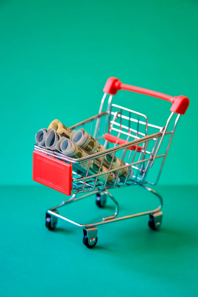 Konzept - zusammengerollte Dollarnoten im Einkaufswagen vor grünem Hintergrund - Foto, Bild